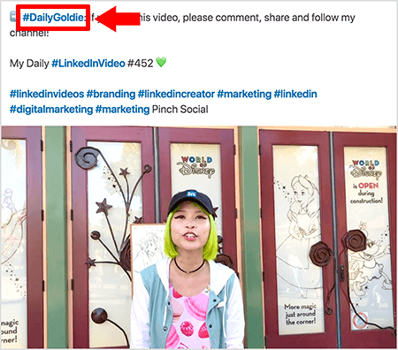 Ovo je snimka zaslona koja ilustrira kako Goldie Chan koristi hashtagove u tekstu svojih video postova na LinkedInu. Crveni oblačići ukazuju na # hashtag #DailyGoldie u tekstu, koji je jedinstven za njezine video postove i pomaže joj u praćenju dijeljenja. Objava uključuje i druge relevantne hashtagove koji pomažu ljudima da pronađu njezin video, uključujući # LinkedInVideo. Na video slici Goldie stoji ispred nekih vrata na izložbi World of Disney. Ona je Azijka sa zelenom kosom. Na sebi ima crnu kapu LinkedIn, crnu ogrlicu od davnina, ružičastu košulju s macaron printom i plavo-bijelu jaknu.